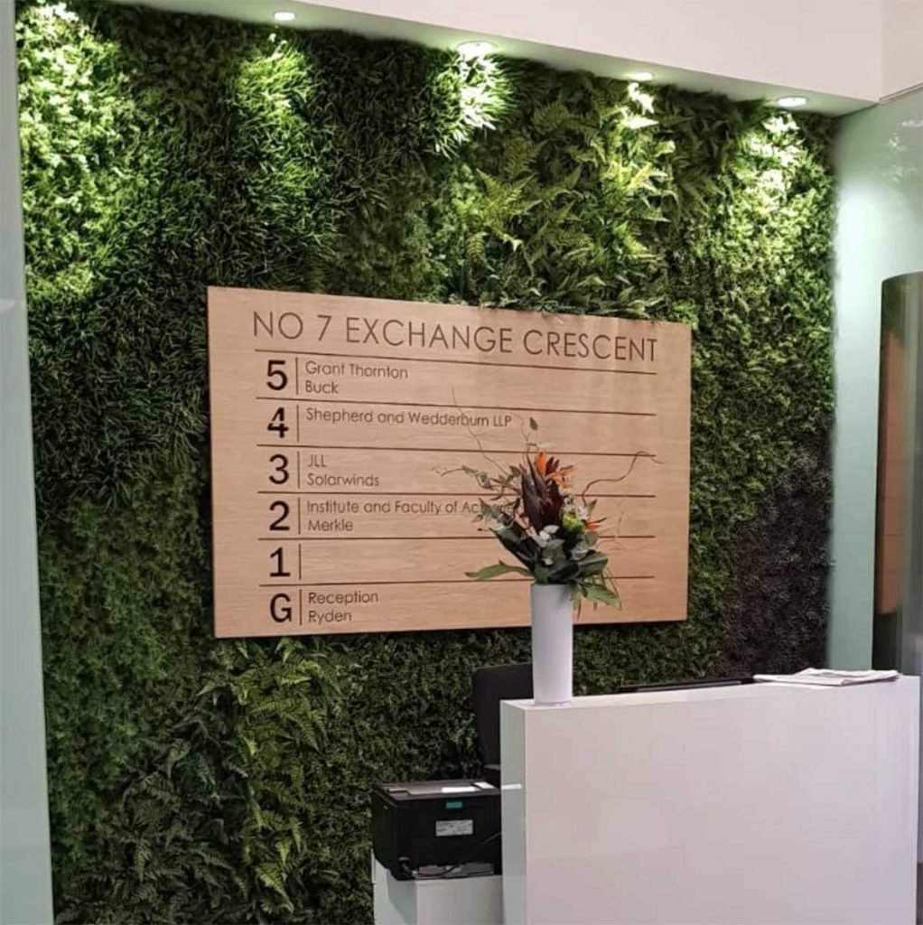 A green wall bursting with greenery lit by spotlights with an inset office directory at a reception space
