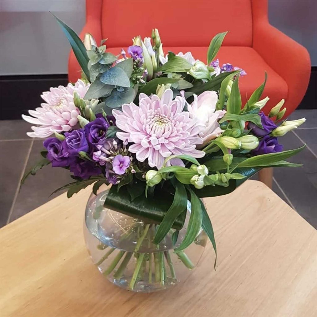 Glass bowl vase filled with freshly cut purple flowers including lisianthus with greenery and leaves